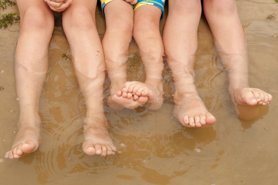 Funny kids in bathing suits sit together on sand with their legs in water. boys are happy on holiday in village together. Summer day, river, swimming in the water. Close-up legs and feet