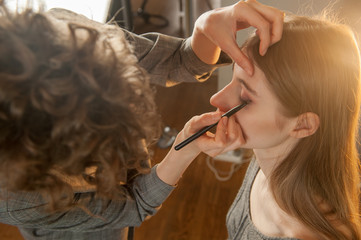 Fototapeta premium Makeup artist and hairdresser are preparing bride before wedding. Hairdresser-stylist with short stylish hairstyle applies makeup, makes false eyelashes, paints lips, curls girl, spectacular lighting