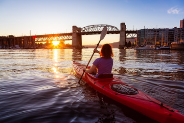 Girl Kayaking in a Modern City during a vibrant golden sunny sunset. Taken in False Creek, Downtown Vancouver, British Columbia, Canada.