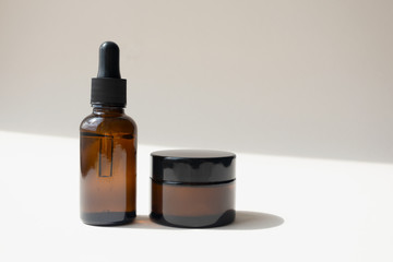 Serum oil essence in brown glass bottle and cream in jar on light beige background with diagonal shadow. Mockup beauty cosmetic products for skincare