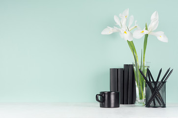 Fresh spring business workplace  - black stationery, books, coffee cup, white flowers in vase on green mint menthe wall and white wood desk.