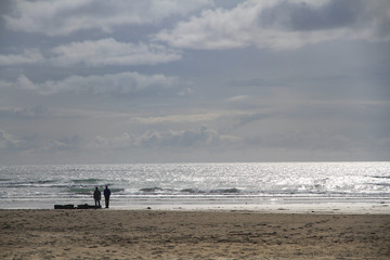 Zwei Personen stehen weit entfernt an einem Strand bei grauen Wolken und sonnenglitzerndem Wasser