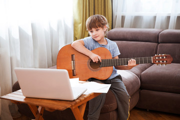 Kid  boy playing  guitar and watching  online lessons  on laptop while practicing at home.  Stay...