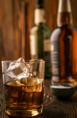 A glass of whiskey with ice, in the background are bottles on a wooden table of a bar counter, shallow depth of field, selective focus. The concept of alcoholic drinks in a roadside bar.