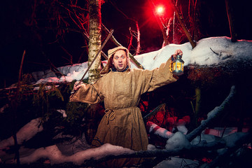 Medieval monk in canvas sackcloth robe with sword and lattern in dark forest and red light of moon...