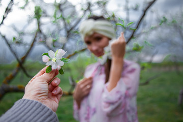 CLose-up of hand holding a flower against a pretty woman dressed pink dress and fashion headdress, walkin in the countryside garden of trees in the nature