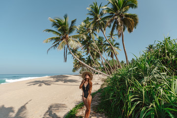 Pretty young woman with closed eyes win  bikini on a background of palm trees. Nice pretty woman holding south straw hat. Green tropical trees and palms background.