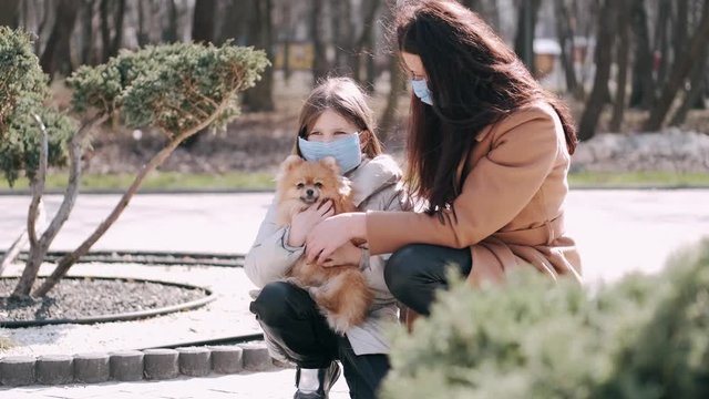 European mother and her daughter are walking with a dog. It's quarantine period so the mother and her child are wearing disposable masks to protect themselves from virus. The parent and chils are