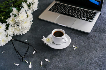on a table is on a black background is a laptop and a coffee mug. near are white chrysanthemums