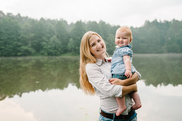Fototapeta premium Mum, little daughter outdoors. Young mother with baby girl walk on beach near lake. Family holiday on pond. Portrait mom with child together in nature. Happy Mothers Day. Close up.