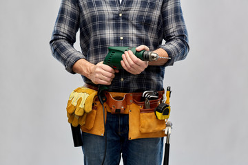 repair, construction and building - male worker or builder with working tools on belt with electric drill or perforator over grey background