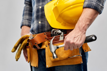 repair, construction and building - male worker or builder with helmet and working tools on belt holding hammer over grey background