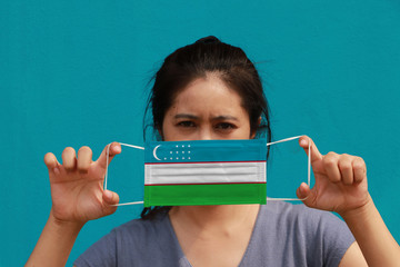 A woman with Uzbekistan flag on hygienic mask in her hand and lifted up the front face on light blue color background. Tiny Particle or virus corona or Covid 19 protection.