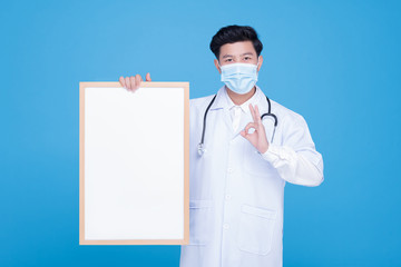 An Asian male doctor wears a surgical mask holding an empty whiteboard horizontally with both hands.
The doctor showing OK sign.