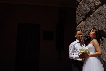 The bride and groom stand near a stone wall on a wedding day in summer and hold a beautiful bouquet. Wedding photo shoot. The bride in a wedding dress. Behind them is a dark background in the shade.