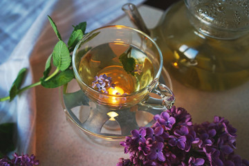 
cup with herbal tea against a teapot with sprigs of mint and lilac flowers. Concept of evening tea drinking