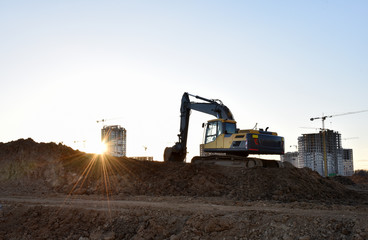 Excavator during earthmoving at construction site on sunset background. Backhoe digs ground for underground sewage and water pipes. Сonstruction machinery for excavating