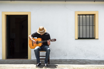 Young man with straw hat playing classical guitar in front of the facade of his white house in the...