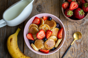 Mini oat pancakes.Mini pancakes with blueberries and strawberries.Pancake porridge, mini pancakes in a bowl with milk and maple syrup.Banana and oatmeal pancakes in a small form.Beautiful breakfast