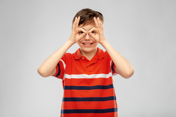 childhood, fashion and people concept - portrait of happy smiling boy in red polo t-shirt looking through finger glasses over grey background