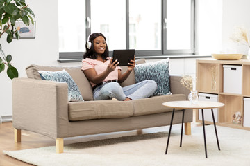 Obraz premium people, technology and leisure concept - happy young african american woman with tablet pc computer and headphones sitting on sofa and listening to music at home