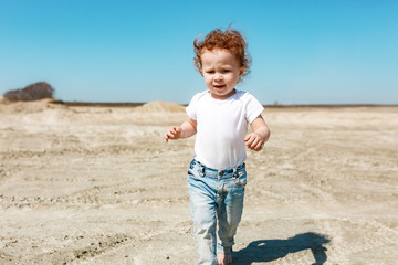 A little girl at the age of 2 walks along the sandy desert in jeans and a white T-shirt. Clear sunny day.