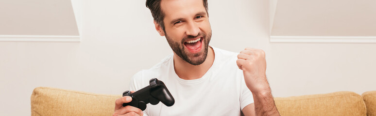 Obraz premium KYIV, UKRAINE - APRIL 14, 2020: Cheerful man holding joystick and showing yeah gesture at home, panoramic shot