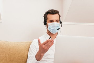 Selective focus of freelancer in medical mask using headset while having video call on laptop at home