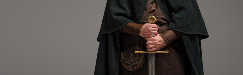 cropped view of medieval Scottish knight in mantel with sword in hands on grey background,...