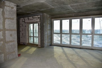 Unfinished interior of apartment under construction with gray concrete wall and double-glazed windows in a residential multi-storey building at a construction site. Housing renovation program