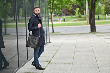 young businessman standing next to a building in coat holding bag 