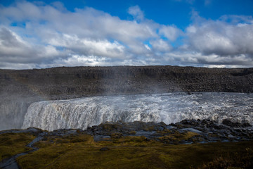 General view of Dettifoss, the most powerful waterfall in Europe. The sky is royal blue with clouds. We can distinguish the mist rising from the surface and a rainbow above