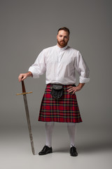 Scottish redhead man in red kilt with sword on grey background