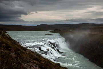 Naklejka na ściany i meble General view of Gullfoss, one of the biggest waterfall in the South West of Iceland. The sky is royal blue with clouds. We can distinguish the mist rising from the surface and a rainbow above