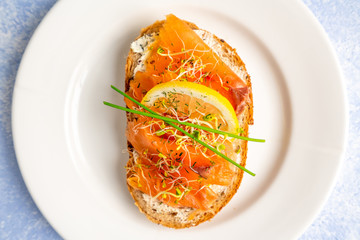 Norwegian smoked salmon with lemon slice, chives and sprouts decoration on a white plate with blue textured backdrop. Food photography with sidelight.