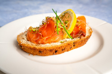 Norwegian smoked salmon with lemon slice, chives and sprouts decoration on a white plate with blue textured backdrop. Food photography with sidelight.