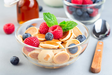 Tiny cereal pancakes served with maple syrup, raspberry, blueberry and milk, in a glass bowl, horizontal