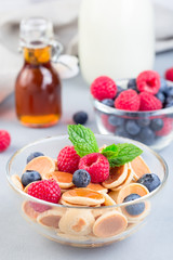 Tiny cereal pancakes served with maple syrup, raspberry, blueberry and milk, in glass bowl, vertical