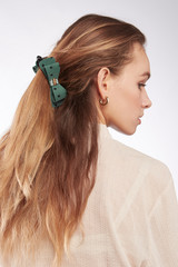 Fototapeta premium A back turned blonde girl with golden earrings is wearing a white blouse. The lady's hair is fixed with a hair clip decorated with a green bow with polka dot print, a golden buckle and a silk ribbon. 
