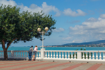 A young couple-a man and a woman, stand on the embankment of the resort of Gelendzhik under a green tree and look out to sea. They are leaning on a white balustrade, with a lantern next to them. 