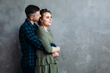 A handsome guy hugs his girlfriend from the back. Happy Cheerful Family. Valentine's Day. Posing against a gray wall.