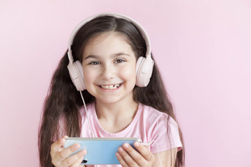 Cute little girl in headphones is using a smartphone, looking at camera and smiling on pastel background