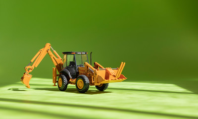 Toy excavator. Model of a wheeled excavator (front loader) with a raised bucket on a green background in the rays of sunlight. Construction equipment. Dreilini/Latvia-04.29.2020.
