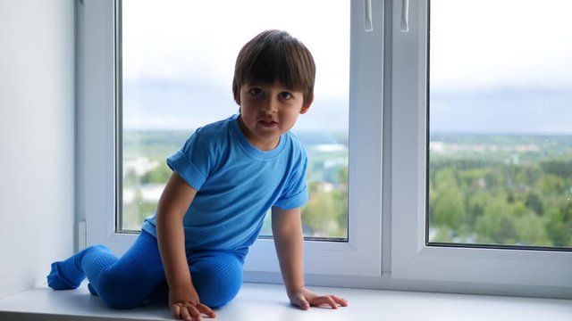 happy child in a blue t-shirt and tights sitting on a windowsill on a high floor in spring
