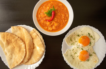 Famous traditional Arabic breakfast - peeled fava beans with chili pepper in white bowl, pita bread and fried eggs on dark wooden background. Top view. Foul Mudammas, egyptian beans, flat bread, eggs