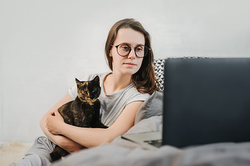 Cute caucasian young woman in eyeglasses relaxing resting on comfortable bed with cute pet cat watching remote education webinar class, movie series on laptop in cozy living room. Social distance
