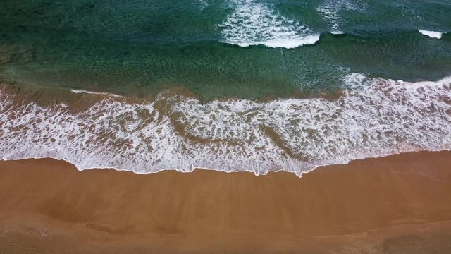 Imagen est&aacute;tica desde mucha altura de la l&iacute;nea de rotura de las olas que nacen, rompen y dejan una estela de espuma hasta llegar a la arena, contraste horizontal entre el mar azul y la arena anaranjad