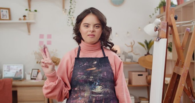 Adorable teen with down syndrome wearing in artists apron showing peace sign while looking to camera. Portrait of cute talanted girl posing while sitting near molbert in her room.