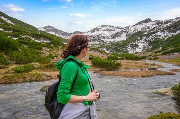 Naklejka premium Redhead Girl with glass looks at the mountains. A woman in green sweatshirt with backpack stands with her back to the camera and looks at the mountain peaks in the snow. Pirin, Bulgaria