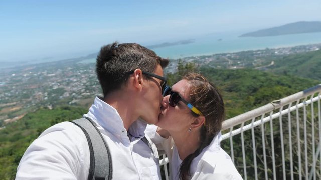 beautiful kissing couple stands on hilltop and makes selfie against pictorial cityscape close up